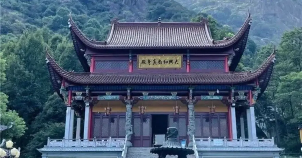 該圖片展示了位於山林間的大興和尚真身殿。主體建築為一座充滿古典韻味的雙層中式廟宇，擁有高聳飛簷與紅褐色屋瓦，正中央懸掛著寫有「大興和尚真身」的金色牌匾。殿前設有灰色石階、精雕蟠龍石柱與一座古樸的香爐，四周環繞著石製護欄。建築背景為連綿起伏、綠意盎然的雄偉山巒，上方襯著藍天與白雲，營造出隱世古剎莊嚴、清幽且與自然和諧共生的佛教聖地氛圍。