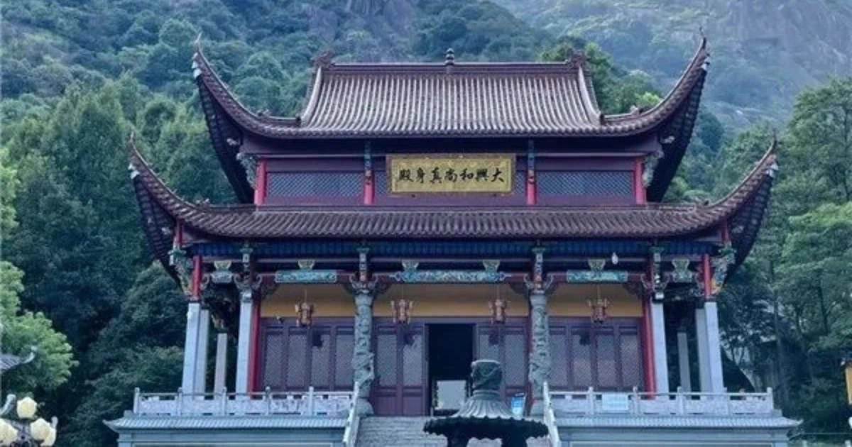 該圖片展示了位於山林間的大興和尚真身殿。主體建築為一座充滿古典韻味的雙層中式廟宇，擁有高聳飛簷與紅褐色屋瓦，正中央懸掛著寫有「大興和尚真身」的金色牌匾。殿前設有灰色石階、精雕蟠龍石柱與一座古樸的香爐，四周環繞著石製護欄。建築背景為連綿起伏、綠意盎然的雄偉山巒，上方襯著藍天與白雲，營造出隱世古剎莊嚴、清幽且與自然和諧共生的佛教聖地氛圍。