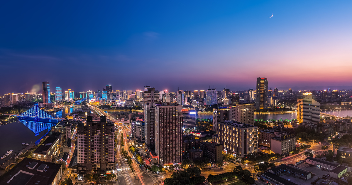 該圖片展示了寧波市繁華的城市夜景與天際線景觀。畫面採取高角度俯瞰視角，流經城市的河道兩岸高樓林立，燈火輝煌，其中左側一座點綴著藍色燈光的現代斜張橋格外醒目，並在水面上投下清晰的倒影。地面上的街道延伸出繁忙的車流燈軌，點亮了整座城市。夜空呈現出由深藍到地平線處橘紅色的漸層暮色，右上角掛著一輪皎潔的細細上弦月，營造出摩登現代卻又不失靜謐的都市夜晚氛圍。